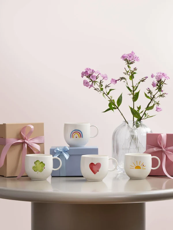 Filiżanki „With love” z motywami takimi jak serce, koniczyna, słońce i tęcza Cztery ozdobione filiżanki „with love” z różnymi motywami, takimi jak serce, koniczyna, słońce i tęcza, dwa pudełka prezentowe i wazon z różowymi kwiatami na okrągłym stole na jasnym tle.