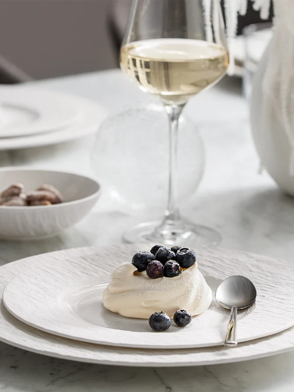 A dessert on a Manufacture plate A dessert on a Manufacture plate garnished with berries, a spoon, a wine glass, and a bowl of nuts on a white tablecloth.