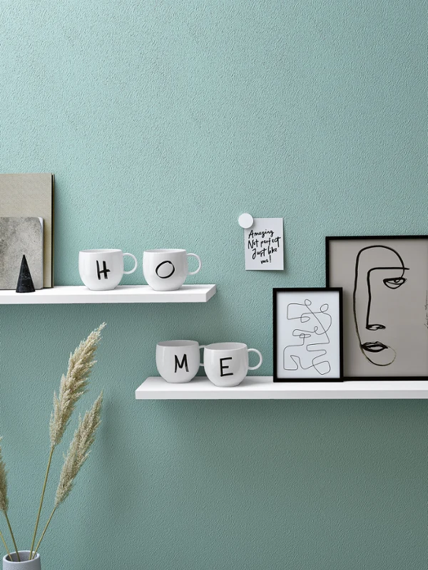 Kubek Letters z literami H, O, M i E na półce Minimalistyczne półki z dekoracyjnymi przedmiotami, w tym kubki z napisem „Home", oprawiony w ramę abstrakcyjny obraz twarzy i roślina doniczkowa.