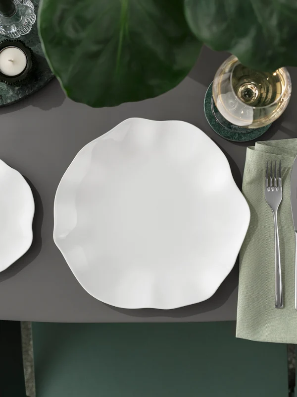 Gedeckter Tisch mit zwei weißen Pura Tellern Gedeckter Tisch mit zwei weißen Pura Tellern, Besteck auf einer Serviette, Brot, grünen Oliven, einem Getränk und einem großen grünen Blatt.