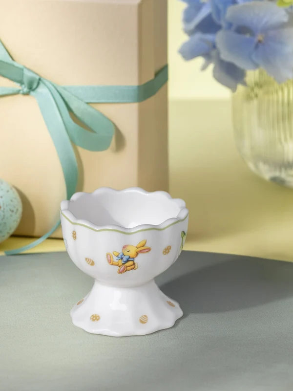 A decorative egg cup from Easter Delight stands on a placemat with two speckled Easter eggs, a wrapped gift, and a vase of flowers in the background.