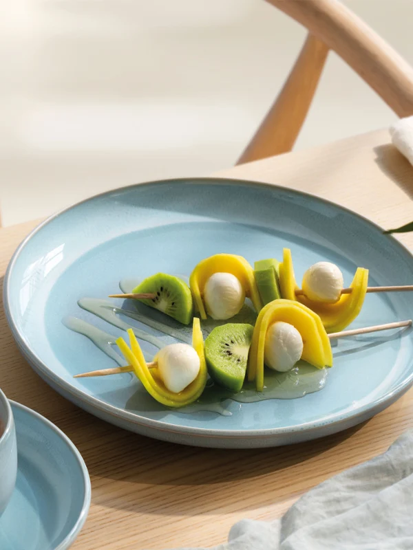 A blue perlemor aqua plate with fruit and mozzarella skewers next to a cup of coffee on a light wooden table.