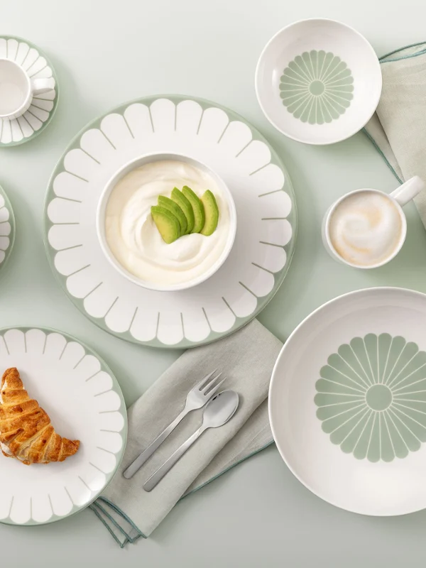 Table setting with Villeroy & Boch Fleur Vert plates, croissant, yogurt, coffee, and white flowers on a light green background.