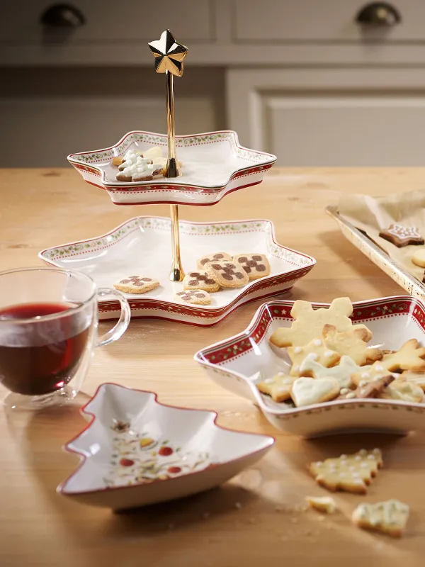 A wooden table with Villeroy & Boch Winter Bakery Delight star-shaped bowls holding decorated biscuits, and a baking tray with festive biscuits on baking paper. There are two glasses with a red drink on the table.