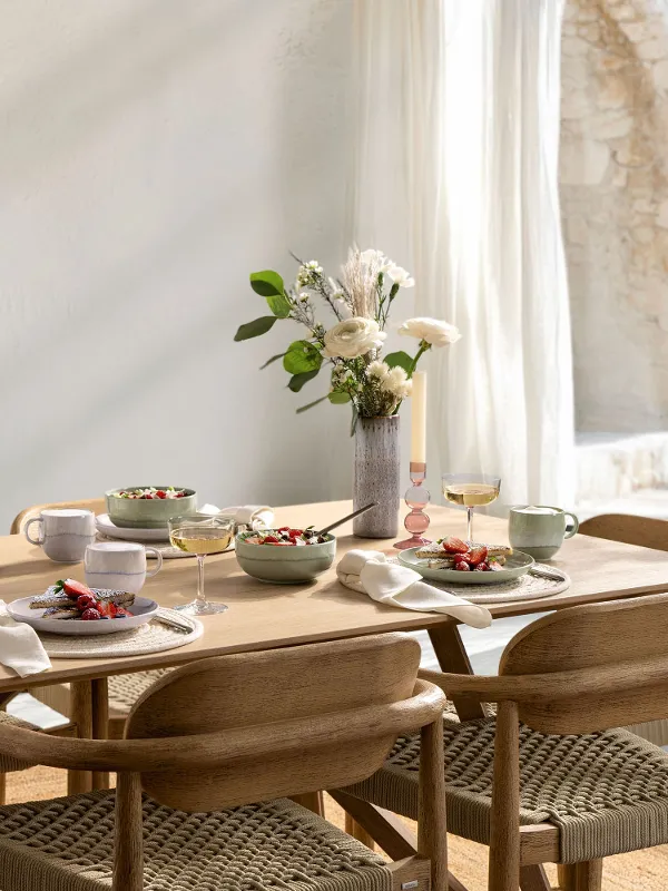 Une table dressée pour six personnes avec des bols, des assiettes et des vases à fleurs dans une pièce ensoleillée dotée de grandes fenêtres et de rideaux clairs.