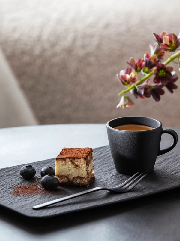 Filiżanka do espresso Manufacture i prostokątny talerz z ciastem Kawałek tiramisu obok czarnej filiżanki do espresso Manufacture, przyozdobiony jagodami na nowoczesnym, prostokątnym talerzu Manufacture z dekoracyjnym wazonem i kwiatami w tle.