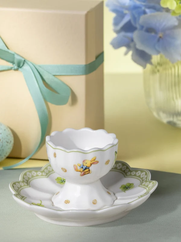 A decorative Easter Delight egg cup and saucer set on a table with a gift box, blue speckled egg, and a vase of flowers in the background.