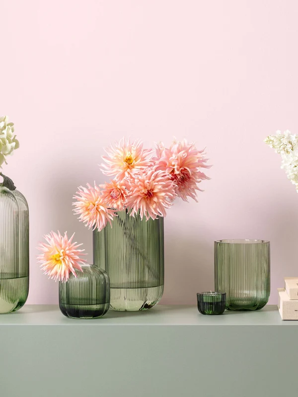 Green Fleur glass vases filled with flowers Green glass vases from Fleur with white and pink flowers arranged on a light green surface in front of a light pink wall.