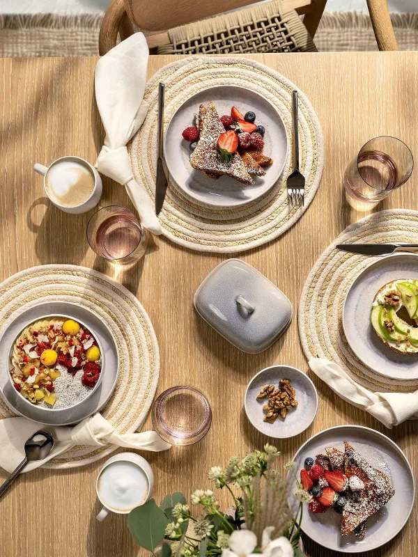 Vista dall’alto di un tavolo in legno accuratamente apparecchiato con pezzi delle collezioni Perlemor Sand e Perlemor Home di Villeroy & Boch: piatto, ciotola per cereali, mug con manico, ciotolina per salse e burriera, accompagnati da bicchieri d’acqua Like Pearl. Le texture naturali, le smaltature morbide e le tonalità equilibrate creano un’atmosfera elegante e rilassata con un tocco artigianale.