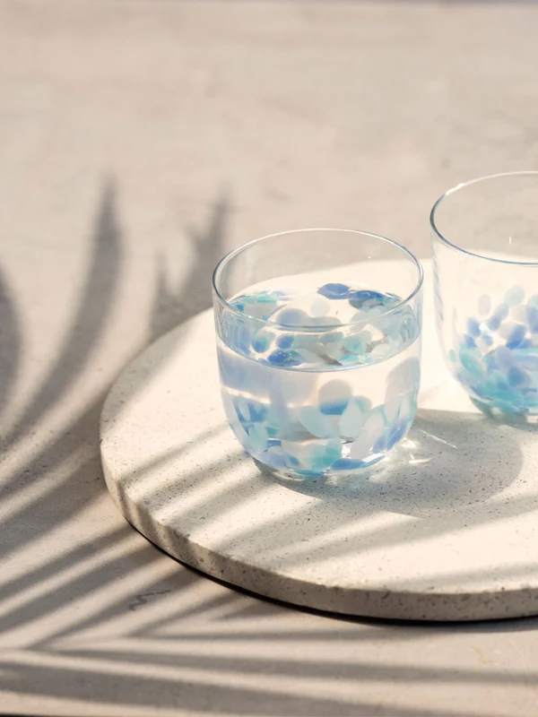 Dos vasos de agua Like Aqua en una bandeja redonda Dos vasos de agua Like Aqua con pétalos azules sobre una bandeja redonda, con sombras de hojas de palmera sobre una superficie clara.