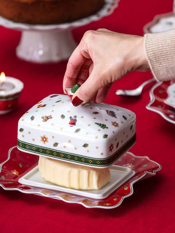 A hand lifting the lid of a festive Villeroy & Boch "Toy's Delight" butter dish on a red tablecloth, surrounded by crockery with a Christmas design.