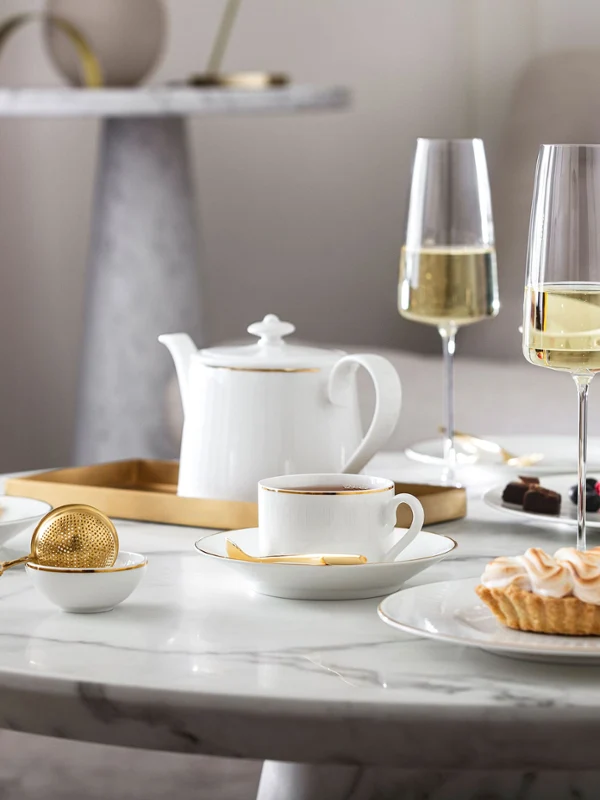 Ein Marmortisch mit weißen Chateau Septfontaines Teetassen, einer Teekanne, MetroChic Champagnergläsern, Desserts und einem goldenen Sieb.