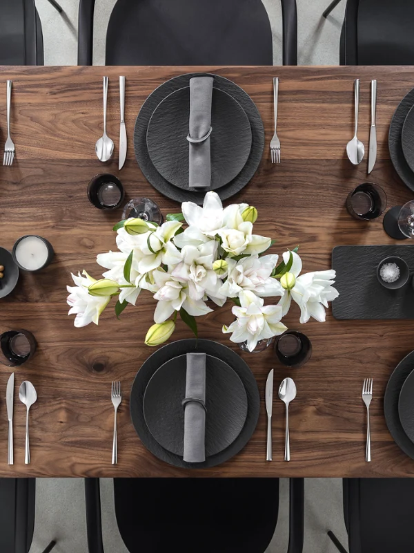 Ein hölzerner Esstisch für sechs Personen mit Tellern von Manufacture, grauen Servietten, Besteck und einer weißen Blumendekoration als Tischschmuck.