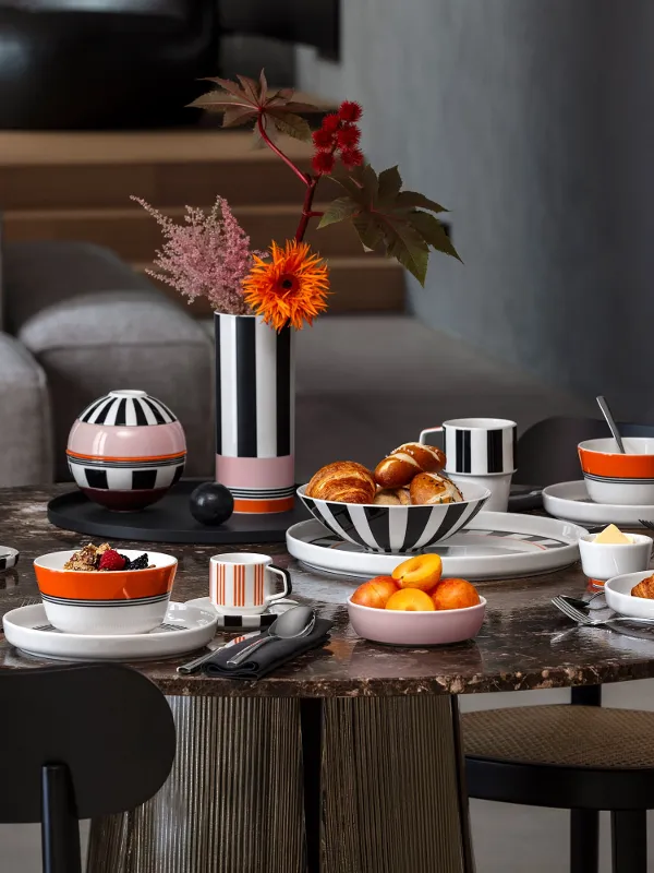 Un tavolo rotondo per la colazione con croissant, frutta, caffè e stoviglie a righe bianche e nere della collezione Memphis di Villeroy & Boch.