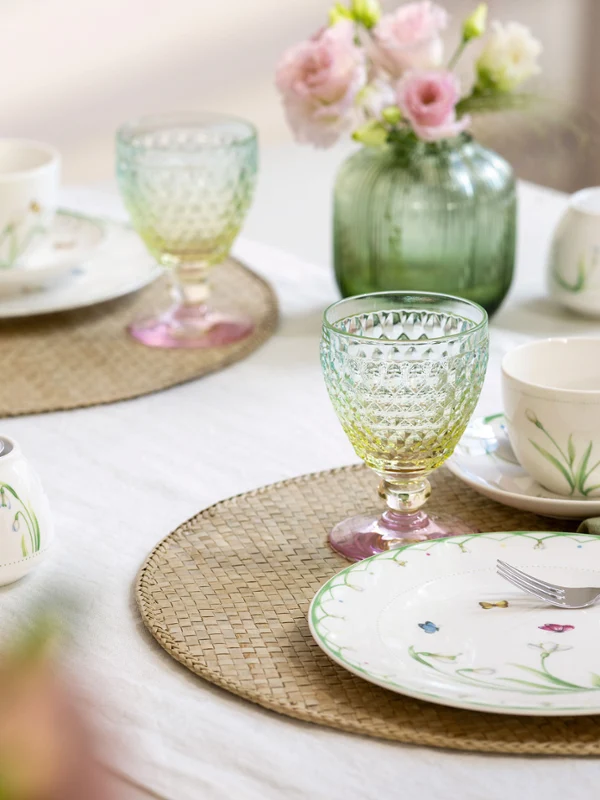 The Colourful Spring set from Villeroy & Boch embellishes any table for two with plates and cups with a floral pattern, bright green and yellow glasses, pink flowers in a vase and beige placemats on a white tablecloth.