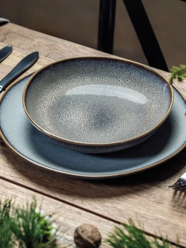 Elegant dining table setting with Lave plates, cutlery and a glass on a wooden table with green foliage.