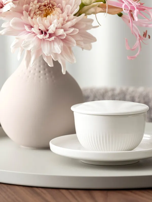 Tasse à café Afina avec soucoupe de Villeroy & Boch, placée devant des fleurs dans un design clair et épuré.