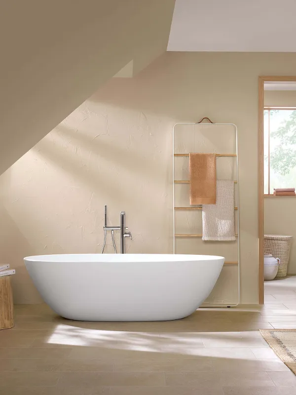 Vasca da bagno free-standing Antao con lavabo da appoggio e rubinetteria in un bagno moderno Bagno con interni moderni, con vasca free-standing, lavabo in legno con piano d'appoggio, WC, specchi rotondi e pareti beige, con luce naturale che entra da una porta aperta.