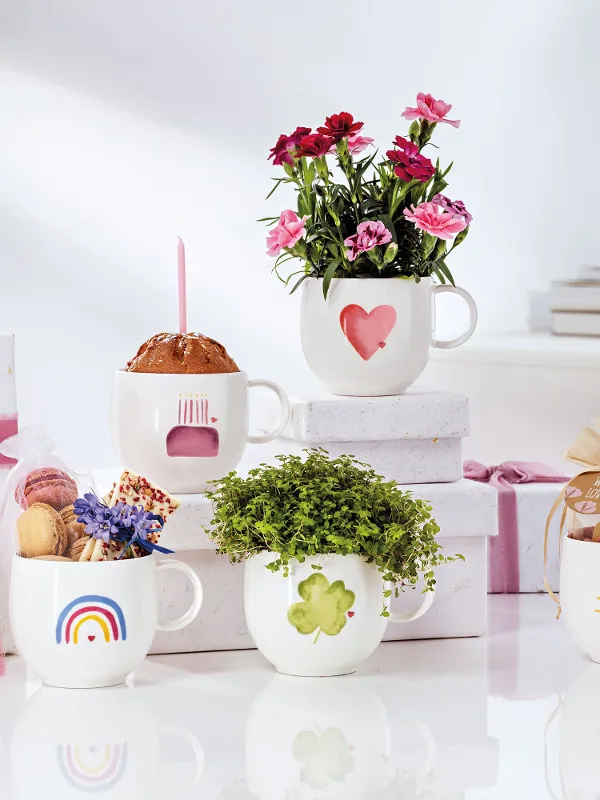 Cinq tasses blanches « With Love » de Villeroy & Boch, chacune avec un motif différent de fleurs, de sucreries et de cadeaux, sont posées sur une surface blanche. En arrière-plan, deux cadeaux emballés avec des rubans roses complètent cette scène enchanteresse.