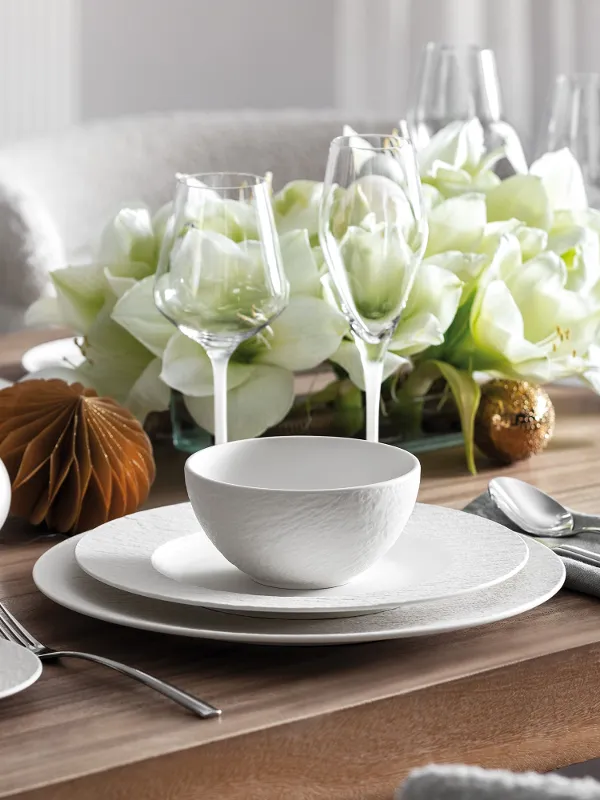 A beautifully laid dining table with the Manufacture Rock Blanc collection from Villeroy & Boch in white, complemented by glassware, silver cutlery and a centrepiece of white flowers and decorative ornaments.