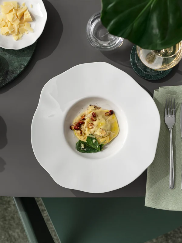 A white Pura pasta plate with pasta and side dishes on a grey table, next to it cutlery, a green napkin and a small bowl of cheese.