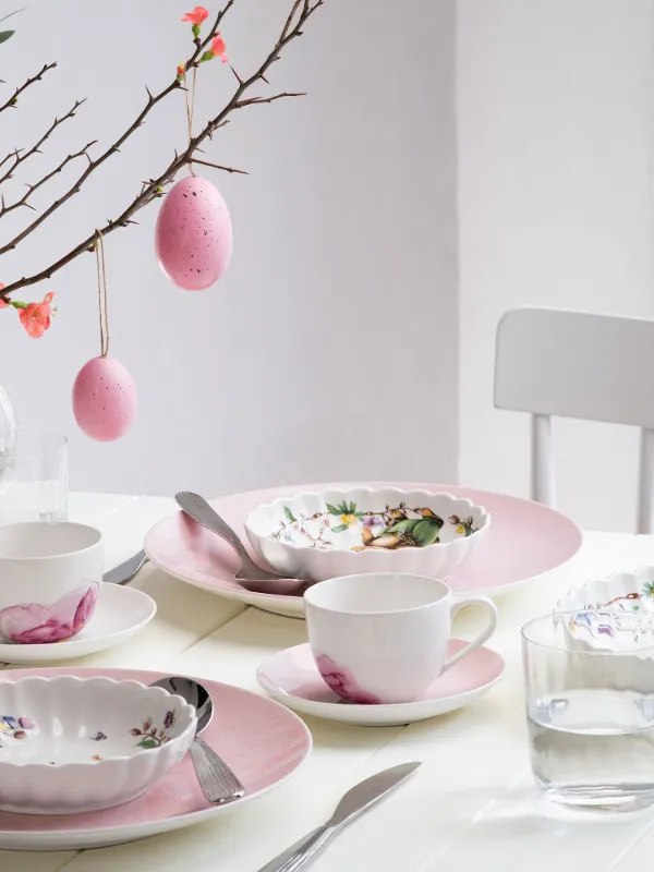 A bright, springtime dining table set with pastel-colored dinnerware from the Rose Garden and Spring Fantasy collections, decorated with floral patterns and Easter eggs hanging from a branch as a centerpiece.