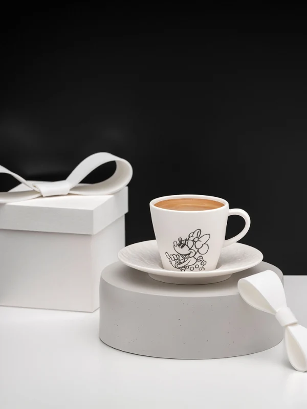 Una taza y un platillo blancos de Mickey Manufacture con motivos negros junto a una caja de regalo. Una taza y un platillo blancos de la marca Manufacture Mickey con motivos negros junto a una caja de regalo y una cinta sobre una superficie minimalista en blanco y negro.
