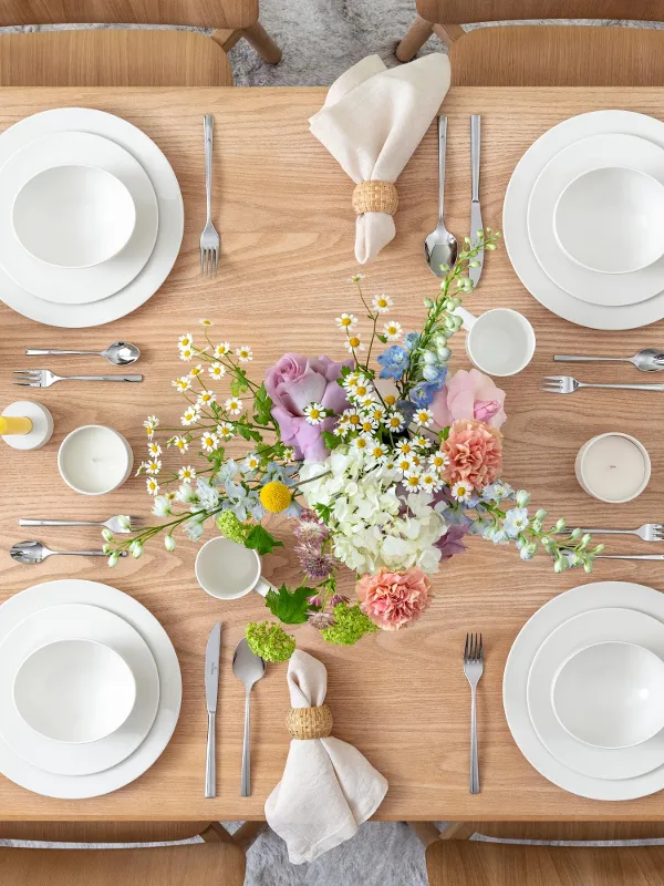 A wooden dining table set for four people with white plates, cups, and cutlery. In the center, there is a floral arrangement with pink and white flowers. Glass jugs are ready, and rolled napkins are placed next to the plates.