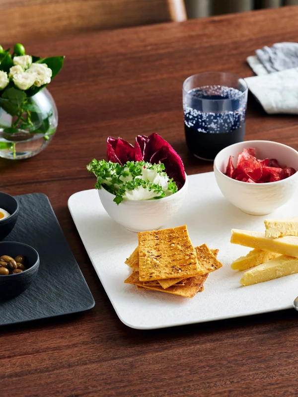 The Manufacture Rock Entertainer Set 6pcs features two black bowls on a black rectangular slate serving plate, alongside two larger white bowls on a large square white slate serving plate, filled with nibbles. Set on a rustic wooden table, complemented with silver cutlery, Manufacture Rock glassware, and Manufacture Rock dinner plate serving prawns.