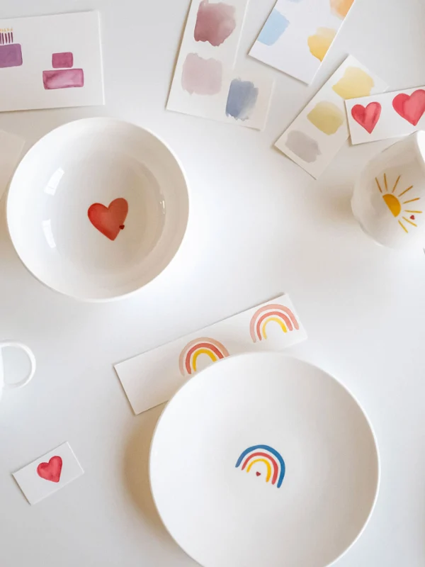 Ein Set weißer Teller und Tassen „With Love“ von Villeroy & Boch, verziert mit lebendigen Herzen, Regenbögen und Baummotiven, elegant arrangiert neben Farbmustern und Musterkarten auf einer makellos weißen Oberfläche.