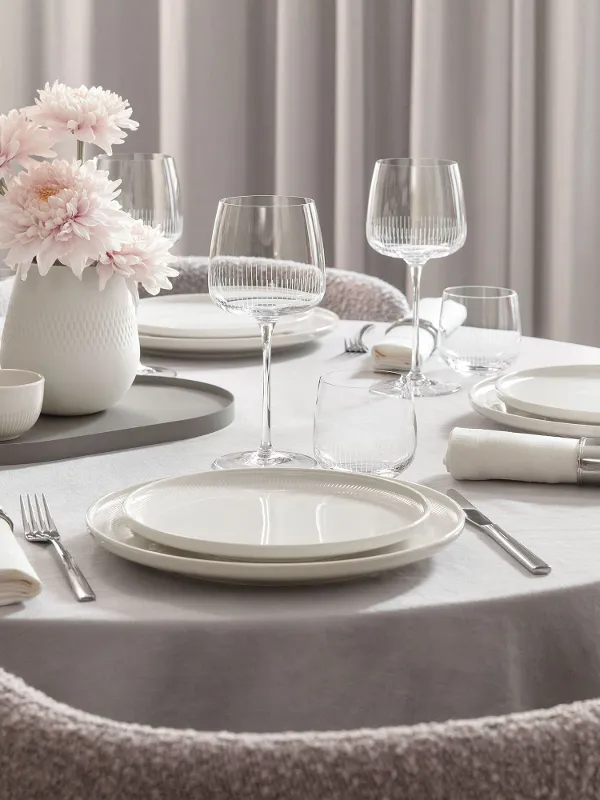 Tavolo da pranzo rotondo con piatti bianchi Afina, bicchieri Afina, posate, tovaglioli e un vaso con fiori rosa.