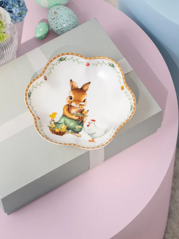 A decorative Annual Easter Edition 2026 plate A gift box with a ribbon sits on a pink table, topped with a decorative Annual Easter Edition 2026 plate featuring a rabbit and flowers.
