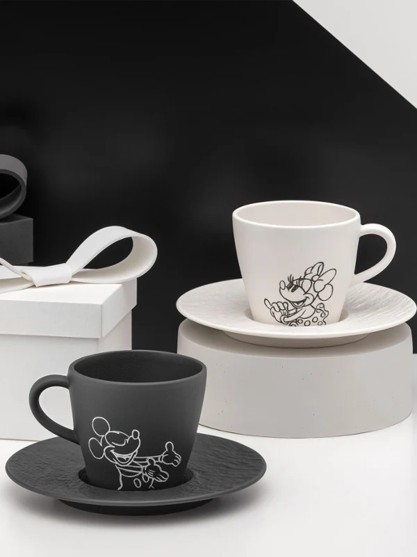 Tazas de té blancas y negras con platillos junto a cajas de regalo Tazas de té blancas y negras con platillos de la colección Manufacture Mickey con cajas de regalo a juego sobre un fondo geométrico en blanco y negro.