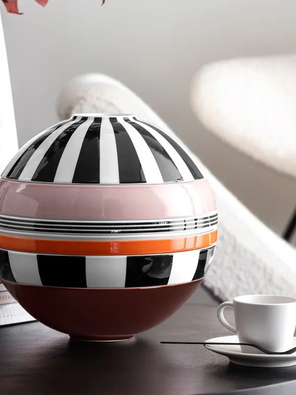 Le modèle Memphis La Boule avec des rayures monochromes et une bande horizontale rose et orange est posé sur une table à côté d’une tasse et d’une sous-tasse blanches, de livres empilés et d’un vase blanc structuré. En arrière-plan se trouve une chaise moderne.