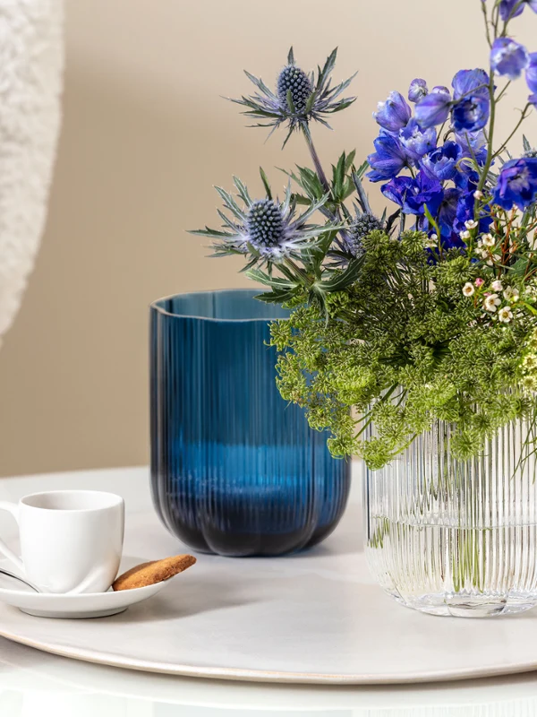 A small white For me cup with a biscuit on a saucer, next to a blue Fleur vase and a transparent Fleur vase A small white For me cup with a biscuit on a saucer, next to a blue Fleur vase and a transparent Fleur vase with blue and green flowers on a round marble surface.