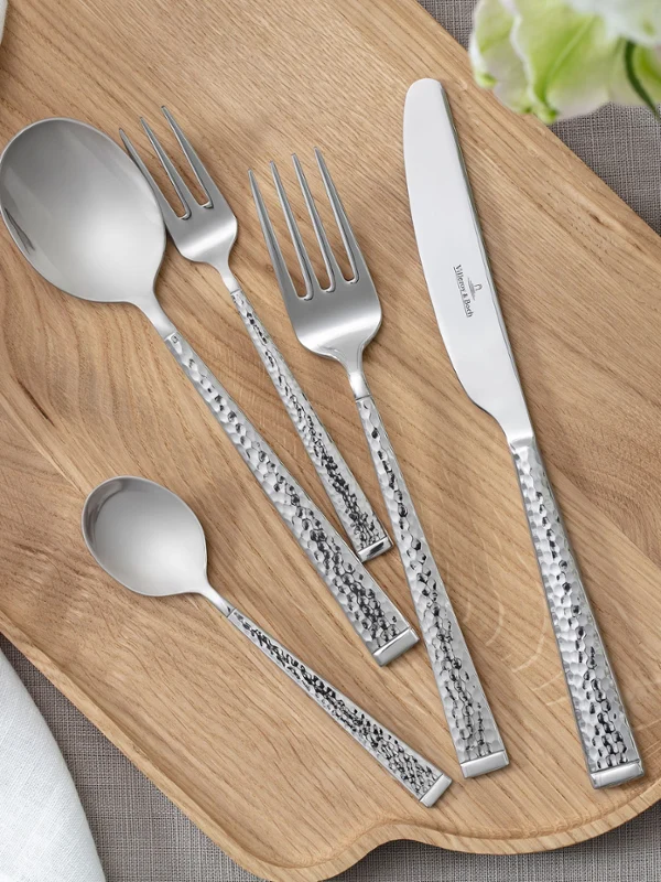 Bovenaanzicht van Blacksmith bestek – soeplepel, tafelvork, tafelmes, koffielepel en dessertvork – netjes gerangschikt op een houten plank. Omlijst door delicate witte bloemdecoratie. Een strakke, natuurlijke en elegante esthetiek.