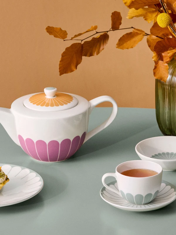 Ein gemütliches Tee-Arrangement mit einer Fleur Teekanne aus Porzellan, Tasse, Untertasse und kleinen Schalen mit pastellfarbenen Blütenblatt-Motiven, abgerundet durch einen Dessertteller mit einer kleinen Tarte und einer grünen Vase mit Herbstlaub.