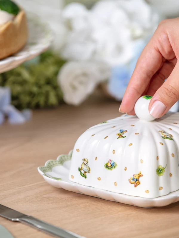 Decorative butter dish from Easter Delight with Easter pattern A hand lifts the lid of a decorative butter dish from Easter Delight on a table set with dessert and a knife.
