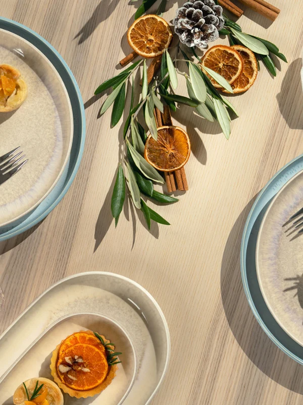 Vista aérea de una mesa de madera clara con un diseño navideño escandinavo con vajilla de porcelana Perlemor de Villeroy & Boch, pequeños pasteles decorados con rodajas de naranja, ramas de canela, hojas de olivo y cubiertos elegantes.