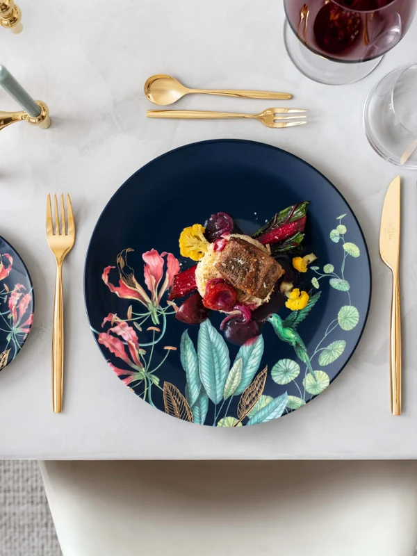 Eleganter Esstisch mit Avarua Tellern und einem Gourmetgericht, dazu goldenes MetroChic Besteck und ein Glas Rotwein.