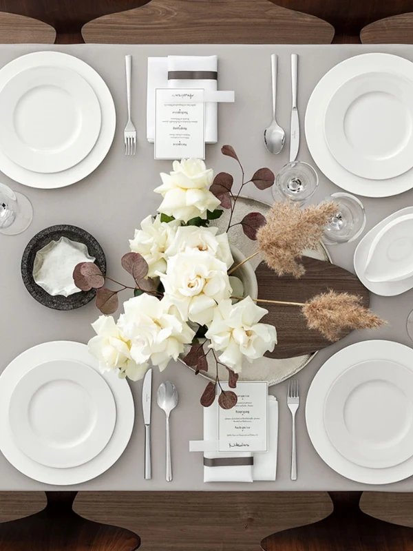 Servicio de mesa con platos blancos Royal de Villeroy & Boch, cristalería con estilo y elegante cubertería de plata sobre un mantel beige. El centro de mesa presenta rosas blancas con toques marrones. Cada asiento viene con un pequeño menú y una servilleta.