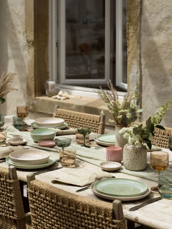 Tavolo da pranzo rustico per esterni con sedie intrecciate, piatti Perlemor color pastello di like. by Villeroy & Boch, bicchieri e centrotavola floreali sullo sfondo di un muro di pietra con finestra.