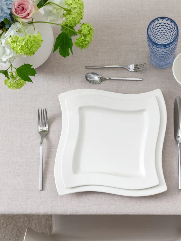 A table set for four with white NewWave plates, salads, cutlery, and napkins. As a centrepiece, flowers in vases are displayed. A clear jug with a drink and scattered candles are also present.