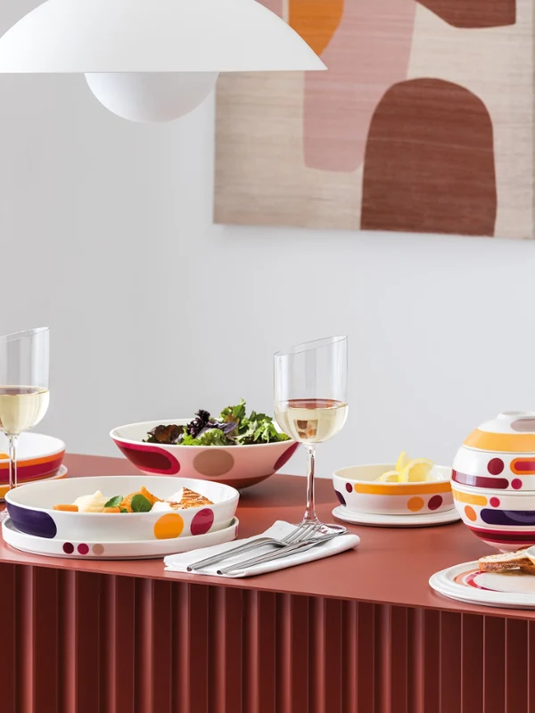 A modern dining table set with colorful patterned dishes, bowls of food, wine glasses, and abstract wall art in the background.