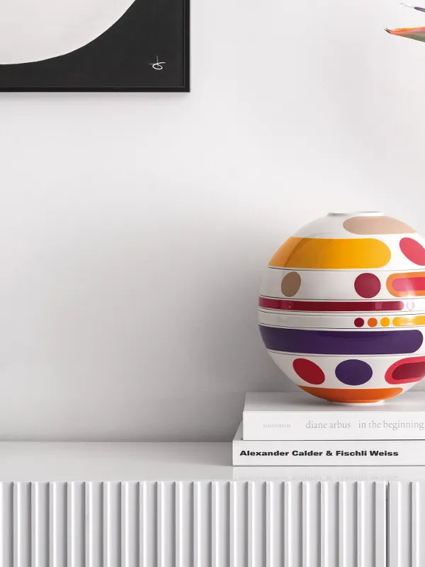 Un modèle La Boule Miami coloré est posé sur des livres à côté de vases blancs et d’oiseaux de paradis sur une étagère blanche.