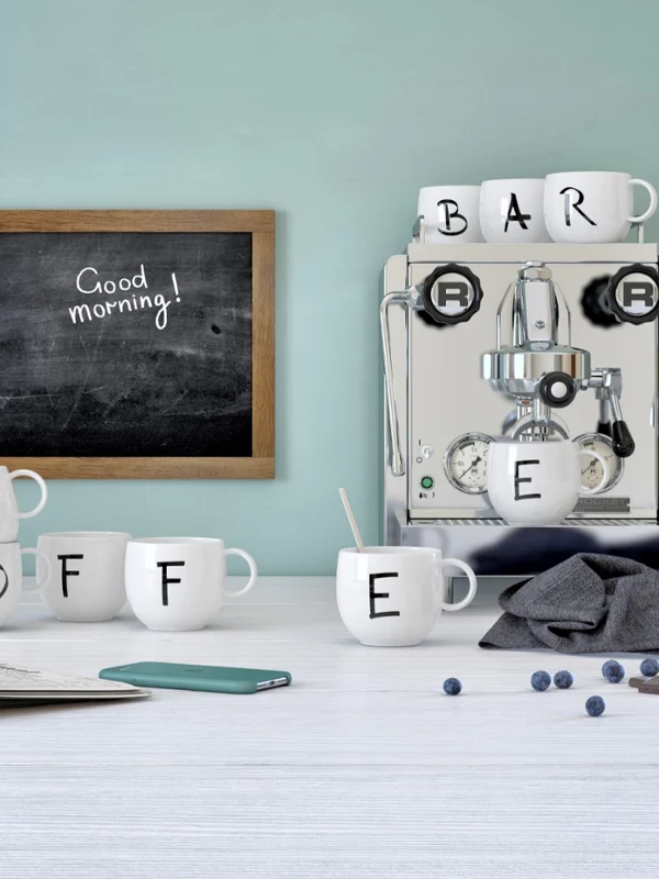 Kubek z kolekcji Letters obok ekspresu do kawy Ekspres do espresso z kubkami Villeroy & Boch „Letters” z napisem „COFFEE” na stole, obok telefonu, gazety, jagód i tablicy z napisem „Dzień dobry!”.