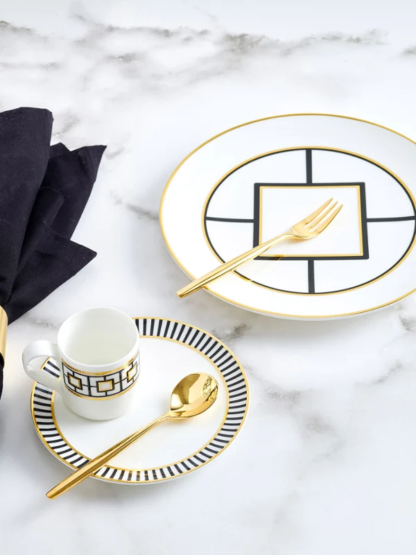 The Metrochic Cafe 4pcs gift set includes a plate, cup, and two pieces of cutlery, all featuring gold accents and a black line design on white porcelain, complemented with a black linen napkin on a white marble table.