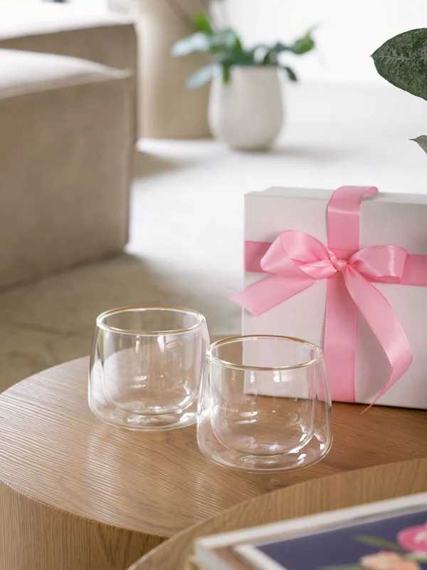 Twee Artesano kopjes van helder glas van Villeroy & Boch staan naast een witte geschenkdoos versierd met een roze strik op een houten tafel, met een beige bank en een plant op de achtergrond.
