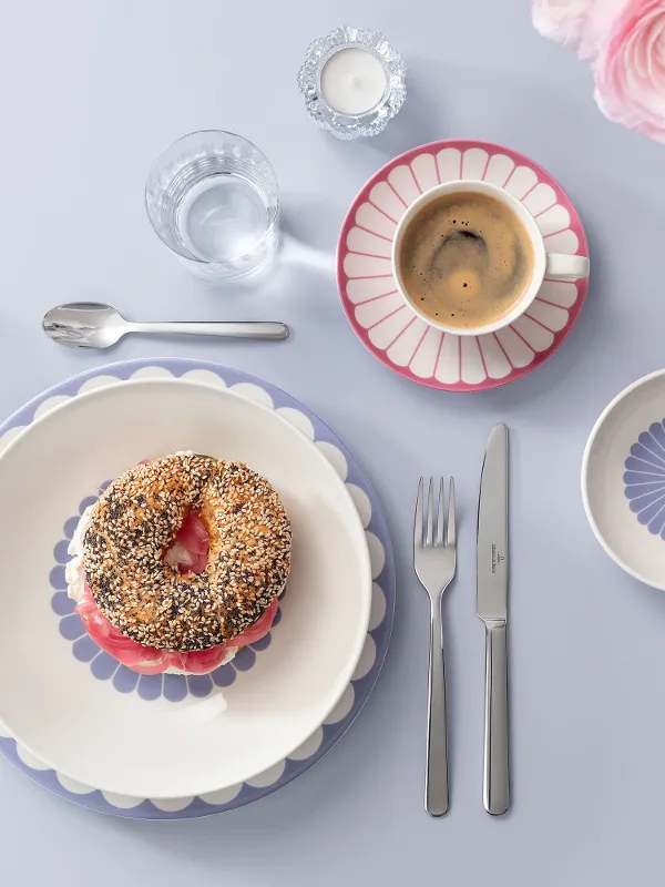 Table setting with a bagel on a decorated plate, surrounded by a coffee cup, small bowls and cutlery on a light blue surface with pink flowers on the side.