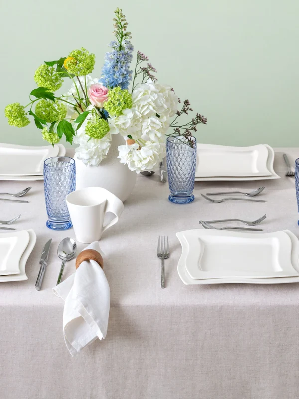 Elegant table setting for four people with white NewWave plates, blue Boston glasses, white mugs, and floral table decorations on a light tablecloth.